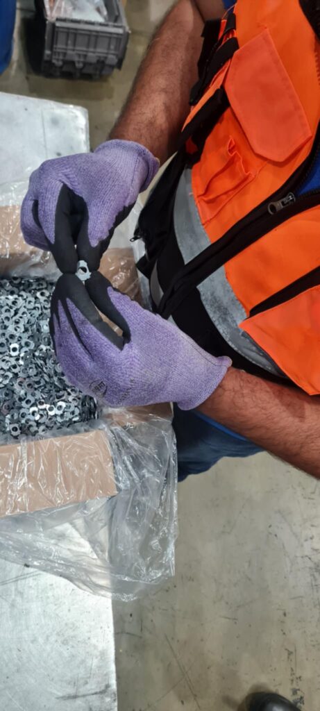 Trabajador con chaleco de seguridad y guantes inspecciona una pieza metálica de una caja llena de componentes, dentro de un área industrial.