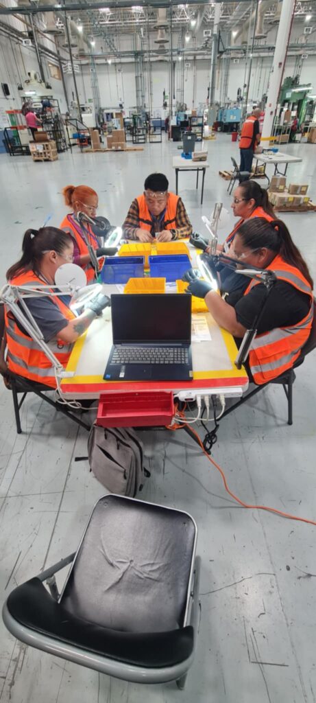 Equipo de trabajo en una línea de inspección dentro de una planta industrial, con trabajadores usando chalecos de seguridad y lupas de aumento para revisar piezas en una mesa equipada con contenedores y una computadora portátil.