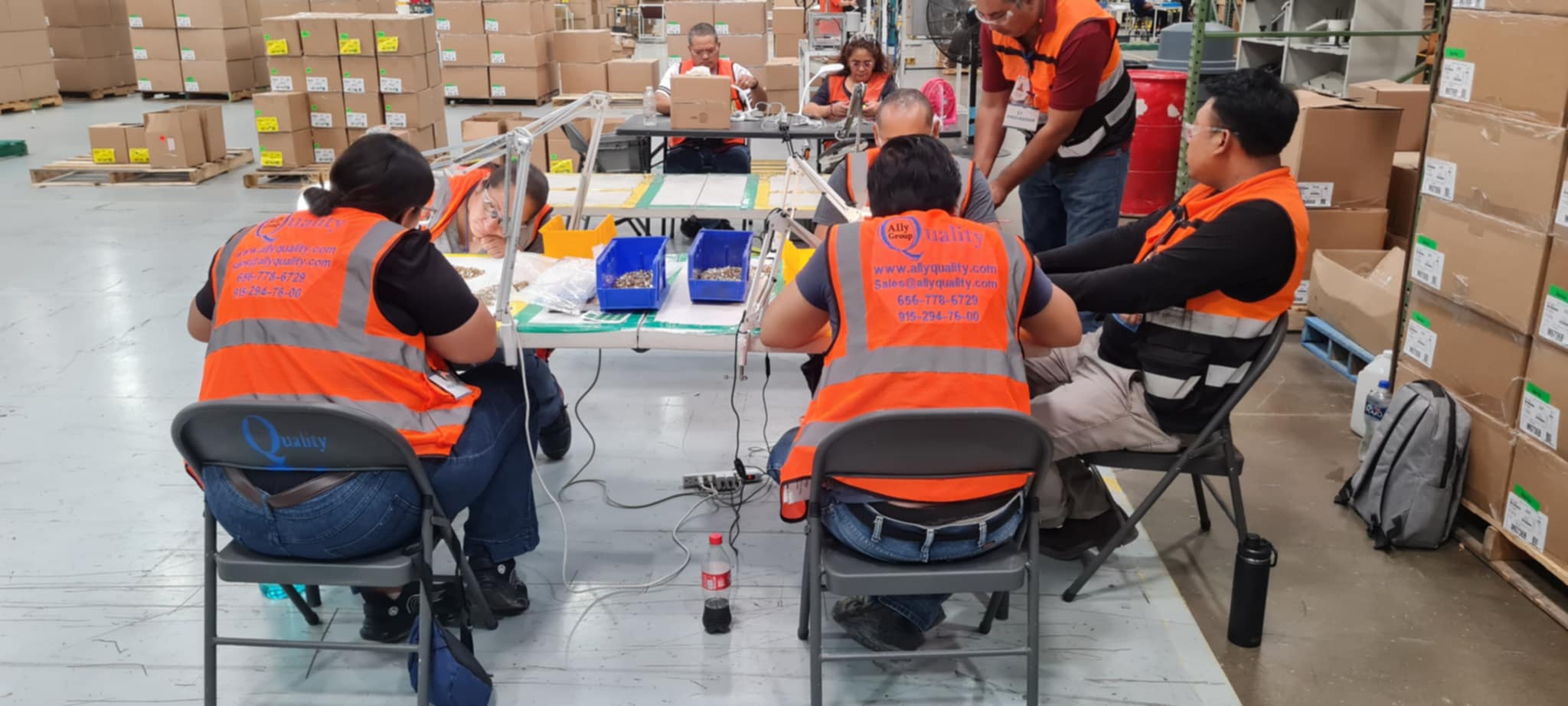 Grupo de trabajadores de Ally Quality Group realizando inspección de piezas en una mesa de trabajo dentro de un almacén, usando chalecos de seguridad y lupas de aumento. Al fondo, más empleados realizan tareas similares entre cajas de materiales.