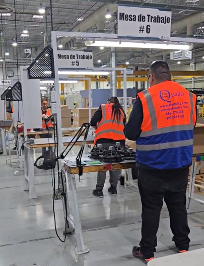 Trabajadores en una planta de manufactura realizando inspección y retrabajo en estaciones de trabajo numeradas. Visten chalecos de seguridad naranjas y azules con el logo de la empresa "Quality". En la imagen se observan mesas organizadas con herramientas, lámparas y componentes en revisión, dentro de un entorno industrial bien iluminado.