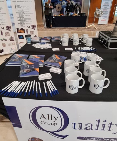 Mesa de exhibición de Ally Quality Group en un evento empresarial, con tazas, bolígrafos, libretas y tarjetas de presentación personalizadas.
