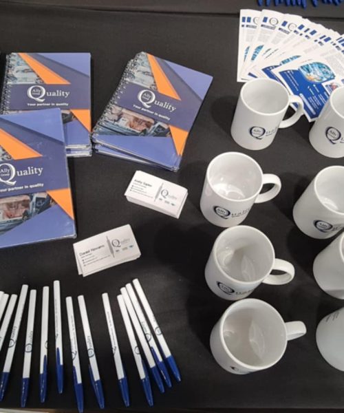Mesa de exhibición con material promocional de Ally Quality Group, incluyendo libretas, bolígrafos, tazas, tarjetas de presentación y folletos informativos sobre los servicios de la empresa. También se observa un termo con el logo de otra compañía y un chaleco de seguridad naranja