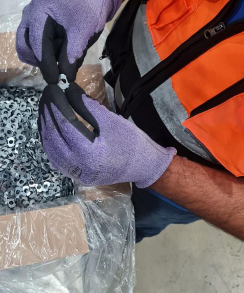 Trabajador con chaleco de seguridad y guantes inspecciona una pieza metálica de una caja llena de componentes, dentro de un área industrial.