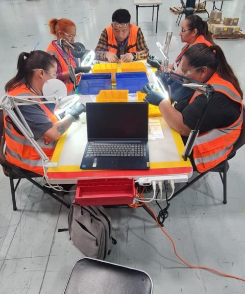 Equipo de trabajo en una línea de inspección dentro de una planta industrial, con trabajadores usando chalecos de seguridad y lupas de aumento para revisar piezas en una mesa equipada con contenedores y una computadora portátil.