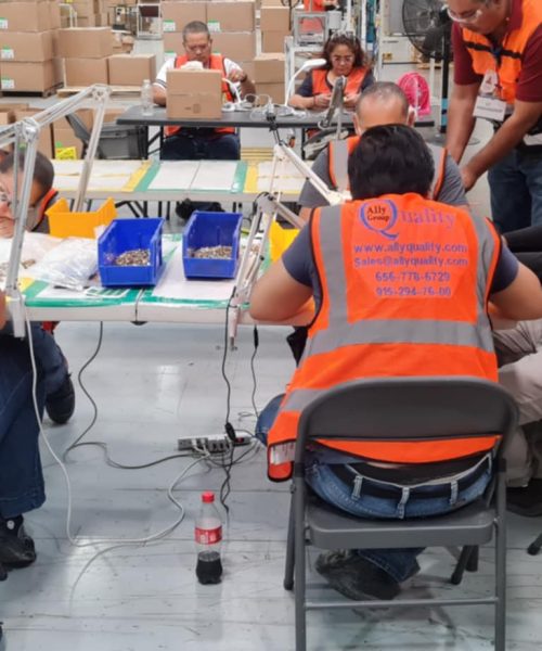 Grupo de trabajadores de Ally Quality Group realizando inspección de piezas en una mesa de trabajo dentro de un almacén, usando chalecos de seguridad y lupas de aumento. Al fondo, más empleados realizan tareas similares entre cajas de materiales.